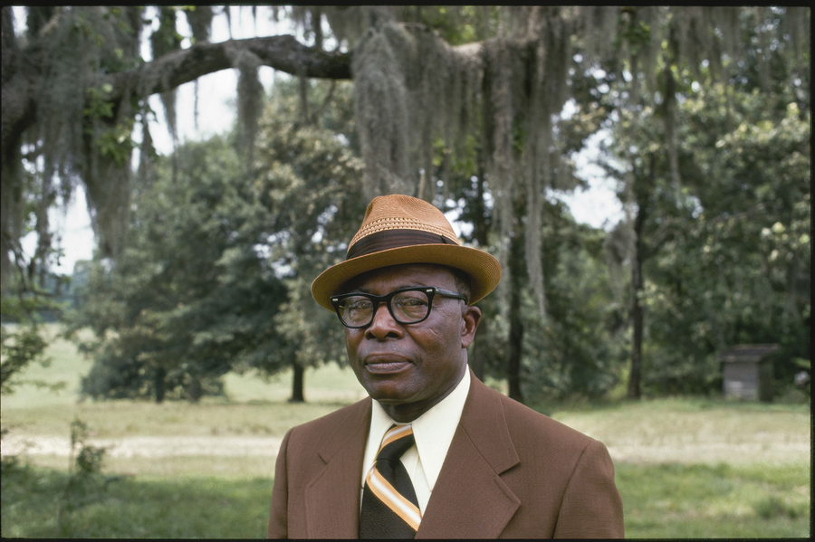 A black man wears a neat brown suit and tan hat in a mid-length portrait photograph. He stares out at the viewer. The background is vibrant green and tree-lined.