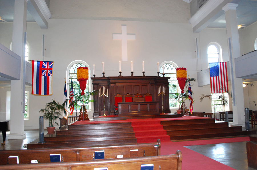 A white church interior includes a wooden altar on a large, stepped platform with a reredos. Lit candles line the reredos and red, white, and blue flags decorate the altar. A simple white cross hangs above the altar.