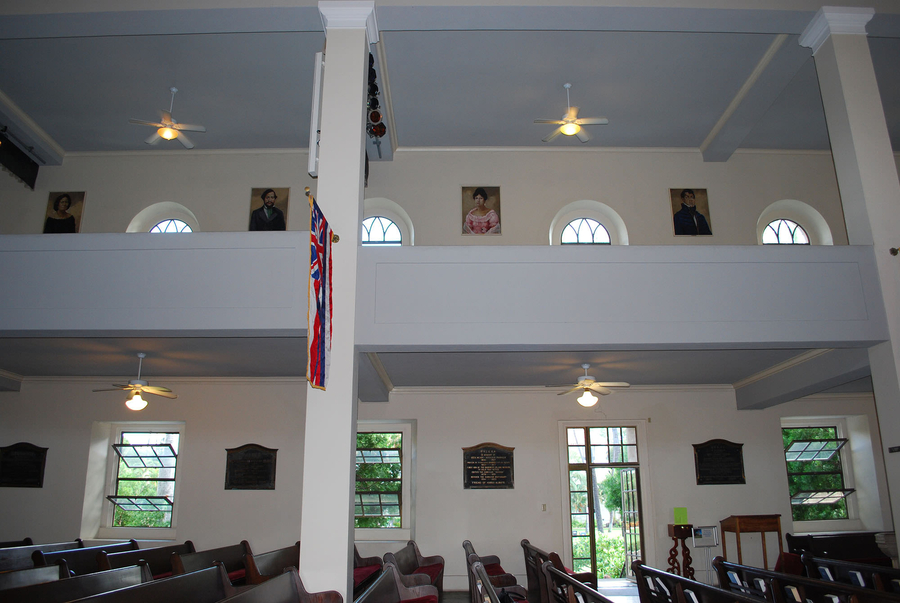A white pew-lined church interior has a balcony that displays painted portraits on its inner walls.