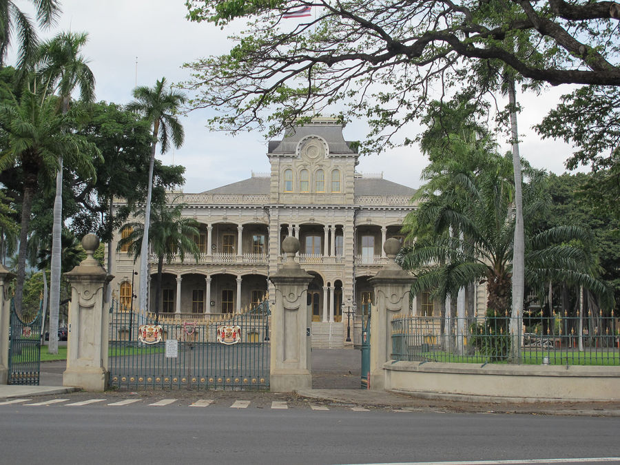 A large gated palace with pale concrete facing has four corner towers and a large, ornamented center tower. Two roofed, open-sided verandas encircle the building on the first and second floors. Palms and other foliage surround the building.