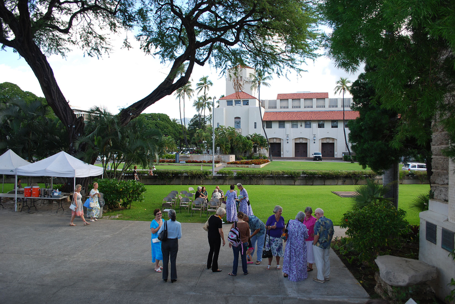 Tourists gather on an open square. It is next to the green lawn in front of a civic hall building.