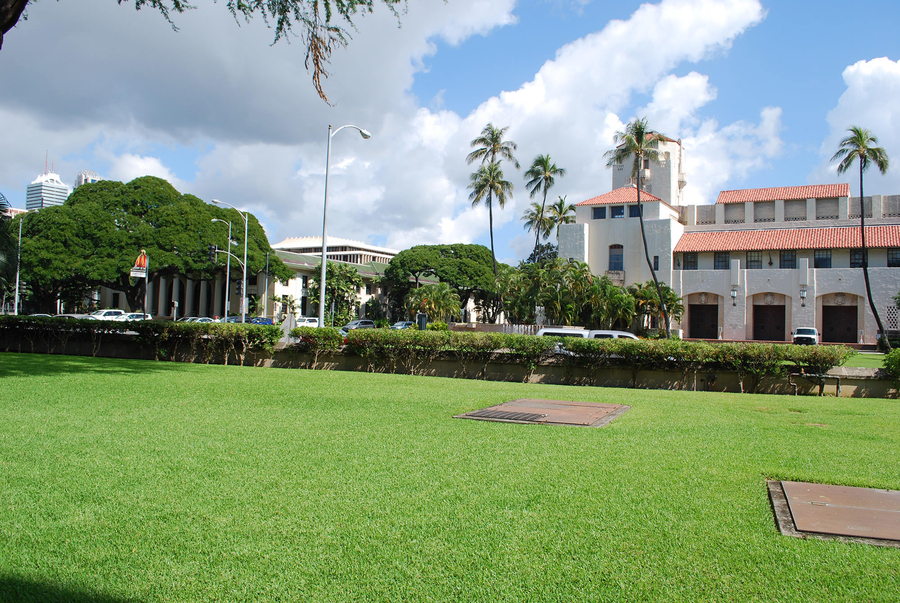 A cluster of white buildings are visible across a manicured lawn. One colonnaded building stands in front of a structure largely blocked by cars and trees. The building across the street has a red tile roof and three large portal entrances.