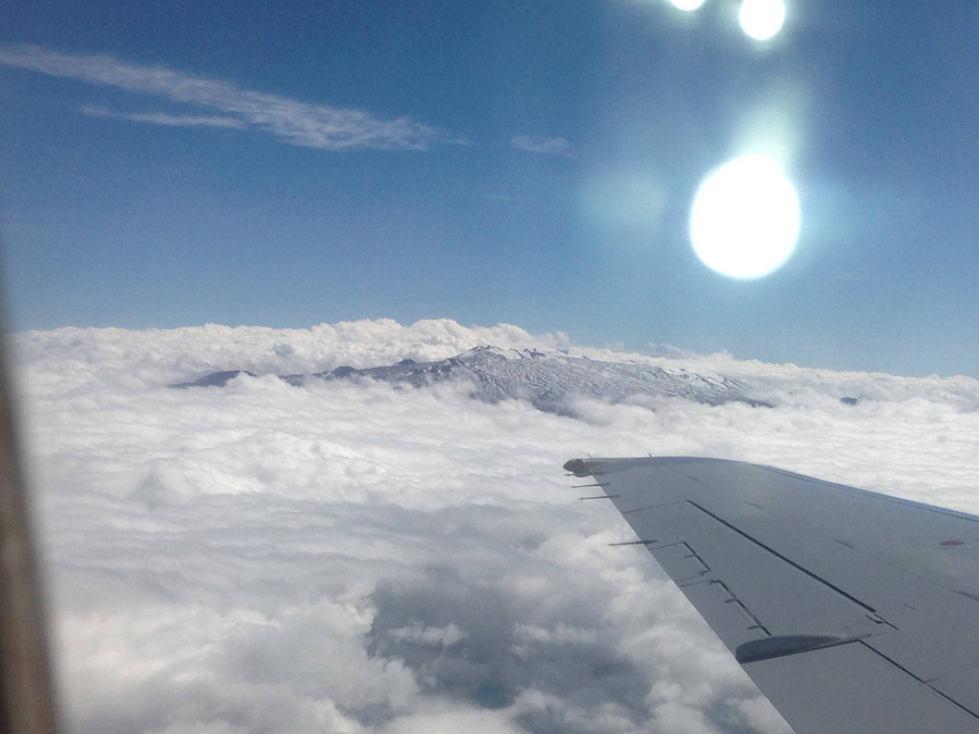 A photo taken from an airplane window captures the plane's wing pointing straight at a mountain summit.  The summit is shrouded in misty clouds.