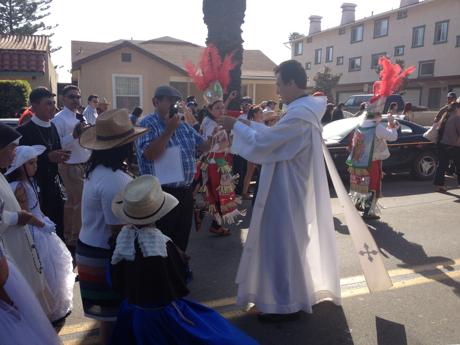 A priest in white vestments interacts with a parading crowd. One man offers him rosary beads to bless while children in straw hats look on. Other paraders wear red feather headdresses as part of colorful, festive costumes.