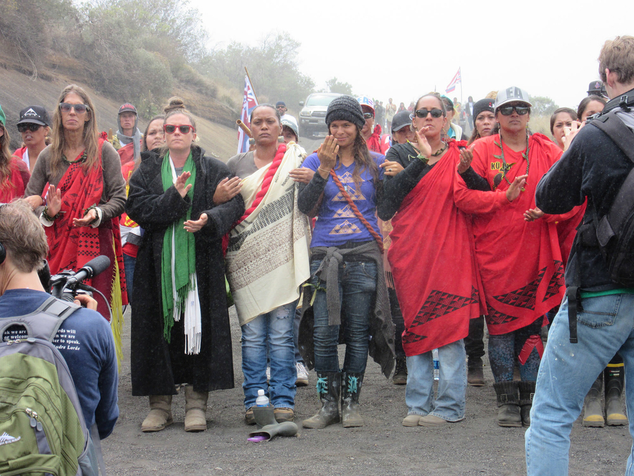 Women link arms and march at the front of a protest. They have serious expressions and gesture with their hands. Some wear red cloaks and men with cameras are visible recording them.