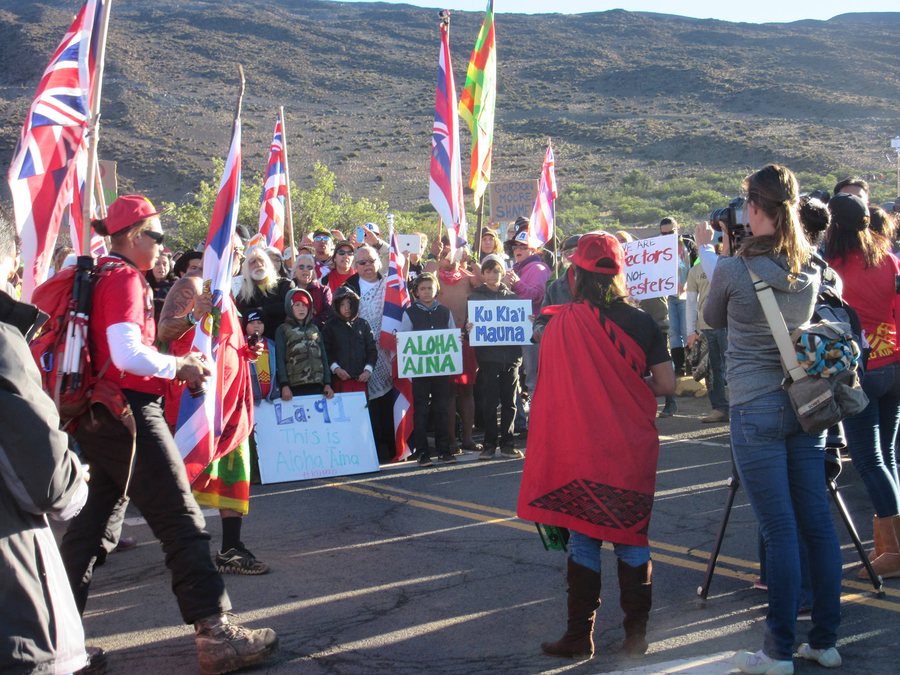 People of many different ages gather on a road in a mountainous landscape. They hold handwritten signs and wave flags. Many wear red. 
