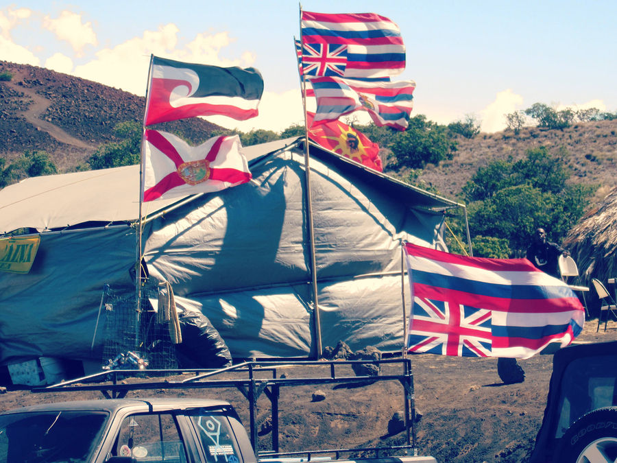Upside-down Hawaiin national flags and other banners hang on flagpoles outside a large, gray billowing tent. 