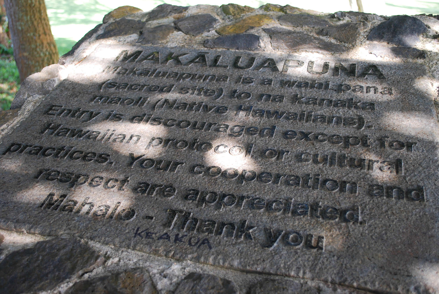 A stone plaque reads, "MAKALUAPUNA Makaluapuna is a wahi pani (sacred site) to na kanaka maoll (Native Hawaiians). Entry is discouraged except for Hawaiian protocol or cultural practices. Your cooperation and respect are appreciated. Mahalo- Thank you." 