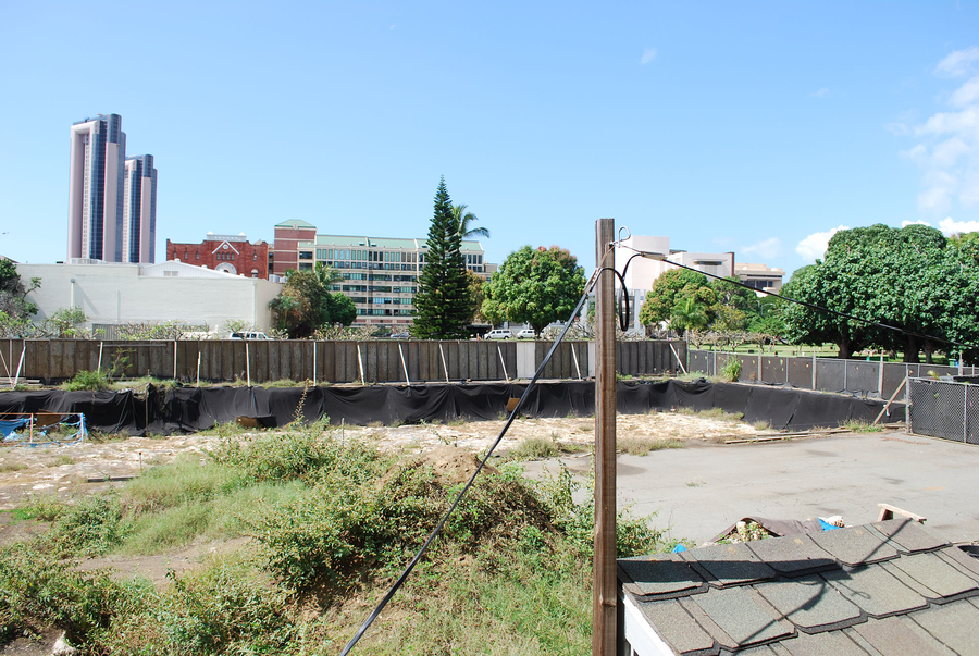 Barricades and fencing enclose a patch of land covered with weeds, asphalt, and tarps. High rise buildings and trees stand in the background.
