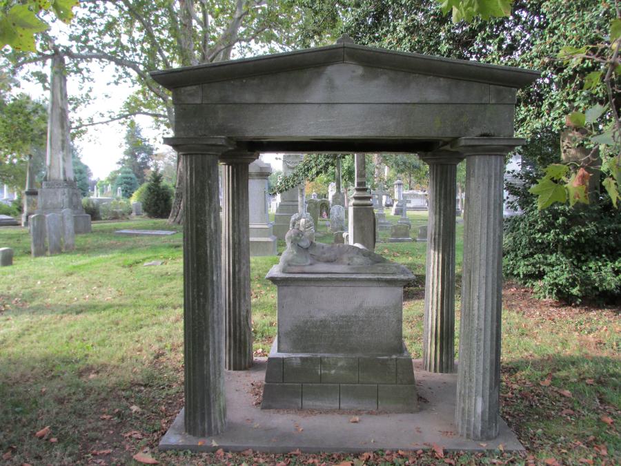 In a graveyard, a stone canopy atop columns shelters a carved child sleeping on a stone podium.