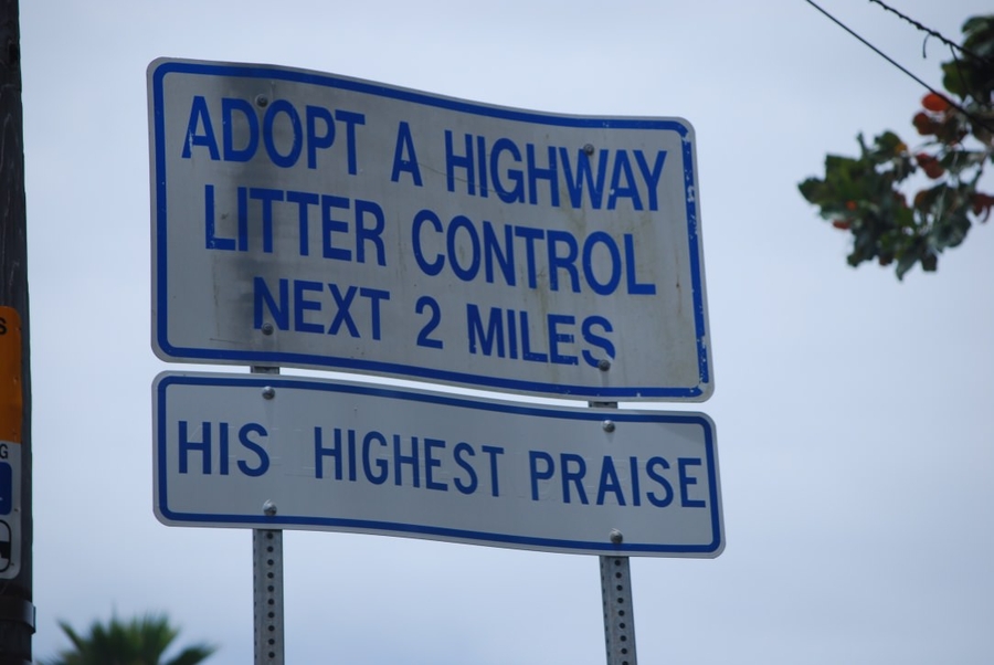 Two white highway signs with blue lettering are affixed one atop the other. The top sign reads, "ADOPT A HIGHWAY LITTER CONTROL." The second reads, "HIS HIGHEST PRAISE."
