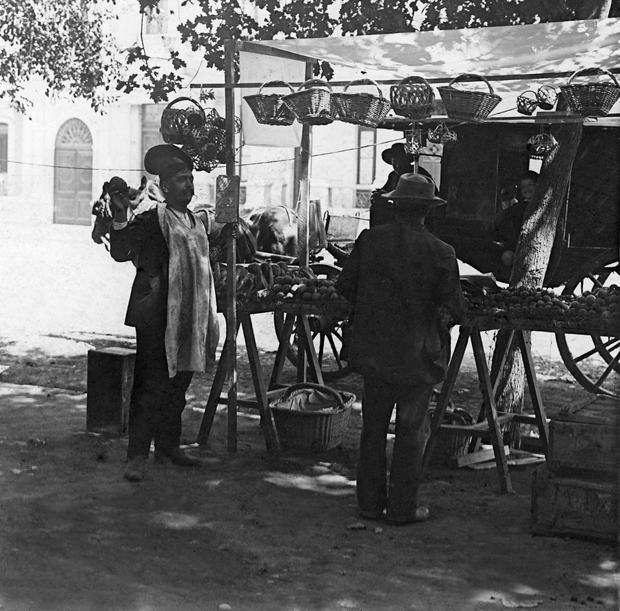 In a black and white photo, two men stand and chat at one of the stalls of an outdoor market. The stall has baskets and fruits for sale.