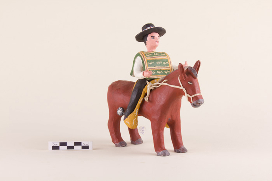 A painted clay figurine depicts a pale-skinned huaso sitting on a brown horse. He has a mustache and wears a black, wide-brimmed hat, yellow chaps, and a green manta. His horse has wide open eyes and has been bridled with a rope.
