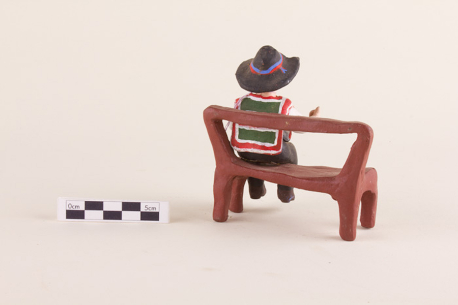 A painted clay figurine depicts a man sitting on a simple brown bench. He wears a brimmed black hat and a green and red poncho.