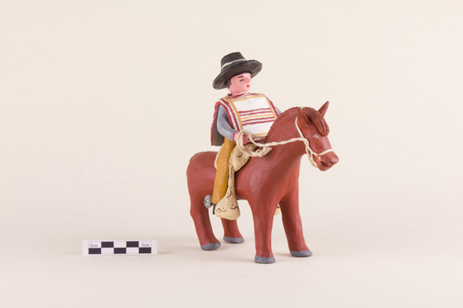 A painted clay figurine depicts a light-skinned huaso sitting on a brown horse bridled with a rope. The man wears a black, wide-brimmed hat, a red and yellow striped manta, and chaps painted with embroidered details.