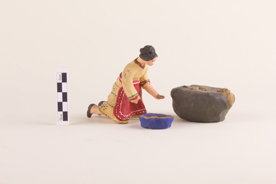 A set of painted clay figurines depicts a woman kneeling before a blue bowl at work kneading bread. She wears a yellow dress and a red apron and holds some dough in her hand. She works next to a larger brown object, which could be an oven for the bread.