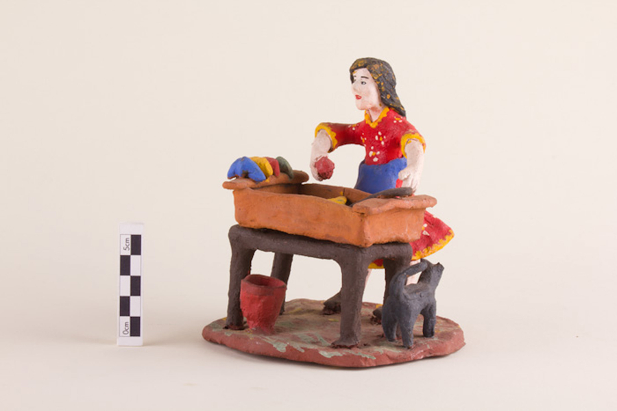 A clay figurine depicts a woman at work washing clothes in an elevated basin. She wears a red dress and a blue apron and grips a red cloth in one hand. More cloths sit on the side of the basin. A small black dog stands by her feet.