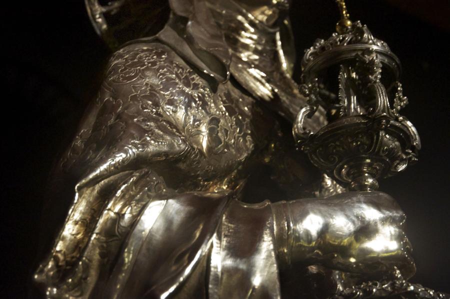 A detail of silver reliquary of Saint Claire focuses on her expertly formed hands. She has slender fingers with incised nails. She grasps a monstance in one hand that glints in the light. 