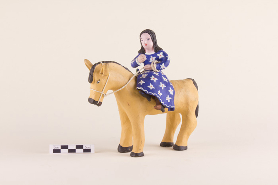 A painted clay figurine represents a woman sitting atop a horse. She is light-skinned with dark hair and wears a purple dress patterned with white flowers. She grips the reins of her honey colored horse.