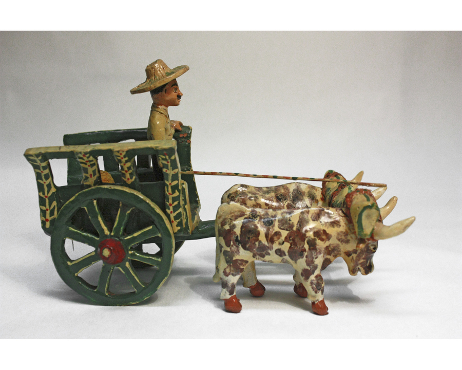 A painted figurine depicts two yoked oxen pulling a green and gold cart. The animals are spotted with brown and have red hooves. A medium-dark skinned man with a mustache and wide-brimmed hat stands in the cart. 