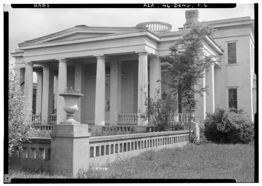 Large columns and square pilasters support the porch of a temple-like mansion. A garden of grass, trees, and decorative balustrades lies in front. It is enclosed by low masonry.