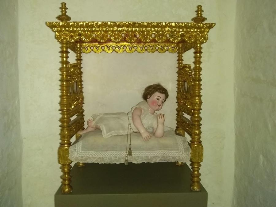 Someone has positioned a light-skinned, wooden sculpture of a child in a small but ornate, golden canopy bed. The child wears a white dress.
