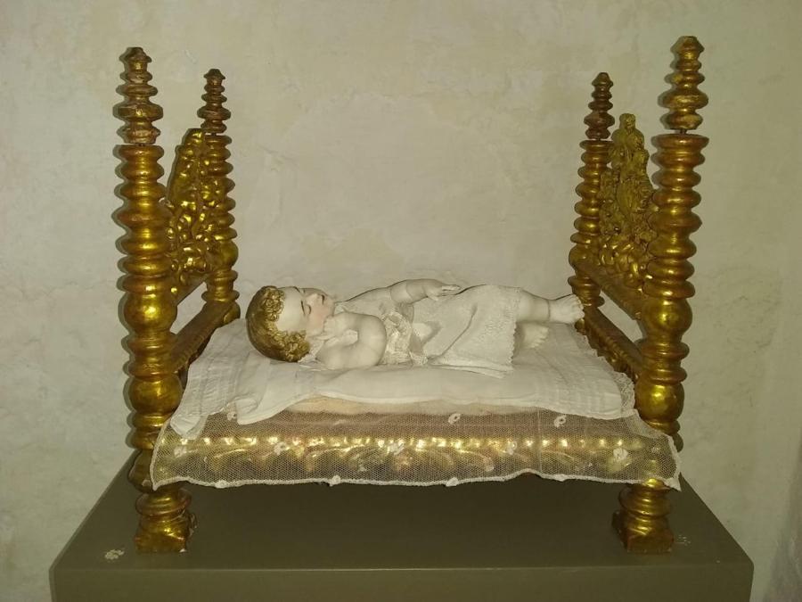 A small golden bed holds a white sculpture of a sleeping child. The child is dressed in a white frock and rests on a gauzy pad.