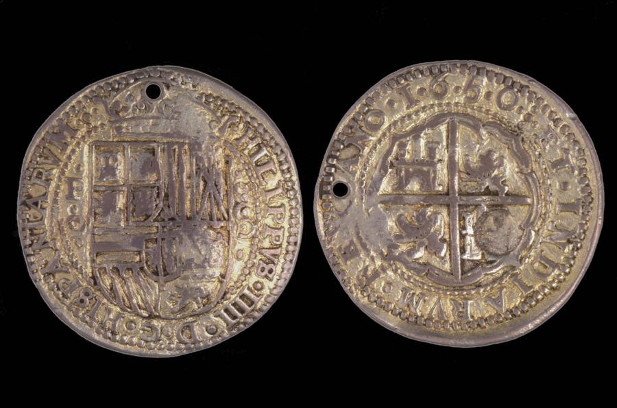 The front and back of a silver Spanish reale are shown side by side. One side depicts a shield surmounted by a crown. The other side has a shield divided into four quadrants with lions and castles.