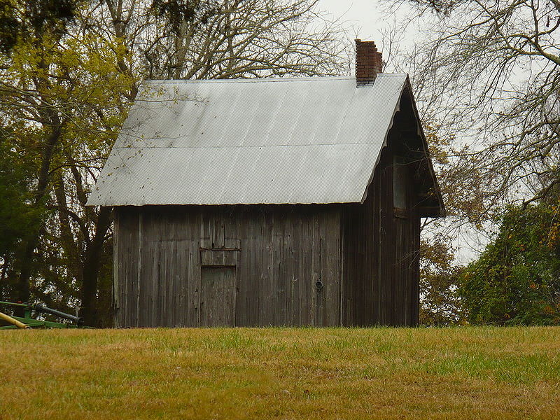 A one-room wooden building has a gable roof and short chimney. It is constructed of dark and deteriorating wood. 