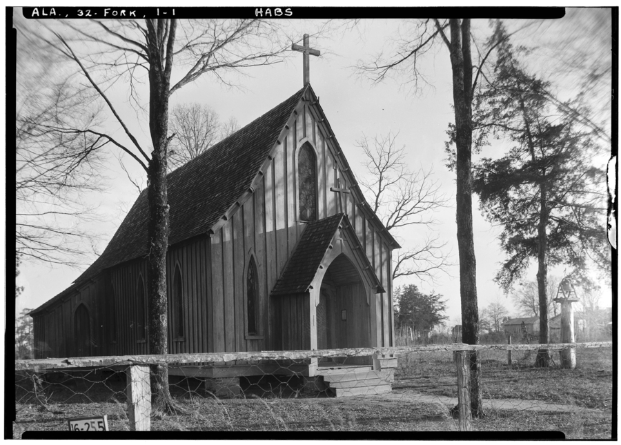 A black and white photograph captures a small church with a curved, cross-topped roof. Vertical battens and pointed arch windows line the church facade.