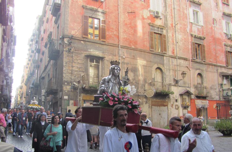 A silver bust of St. Patricia is processed through the street on a litter held aloft by Italian men of different ages. People of different ages and genders process behind them. More litters with busts are also visible among the crowd.