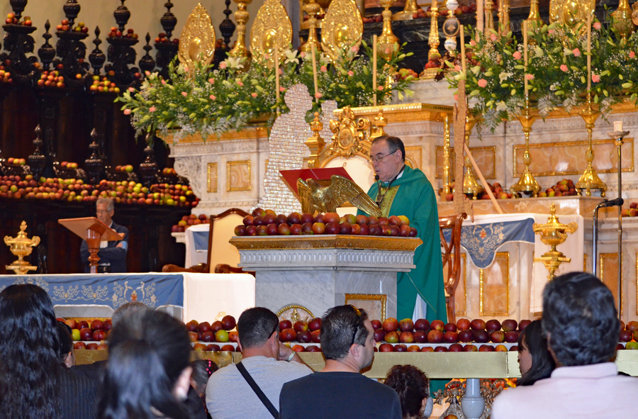 Catholic priest standing at a lectern surrounded by piles of apples