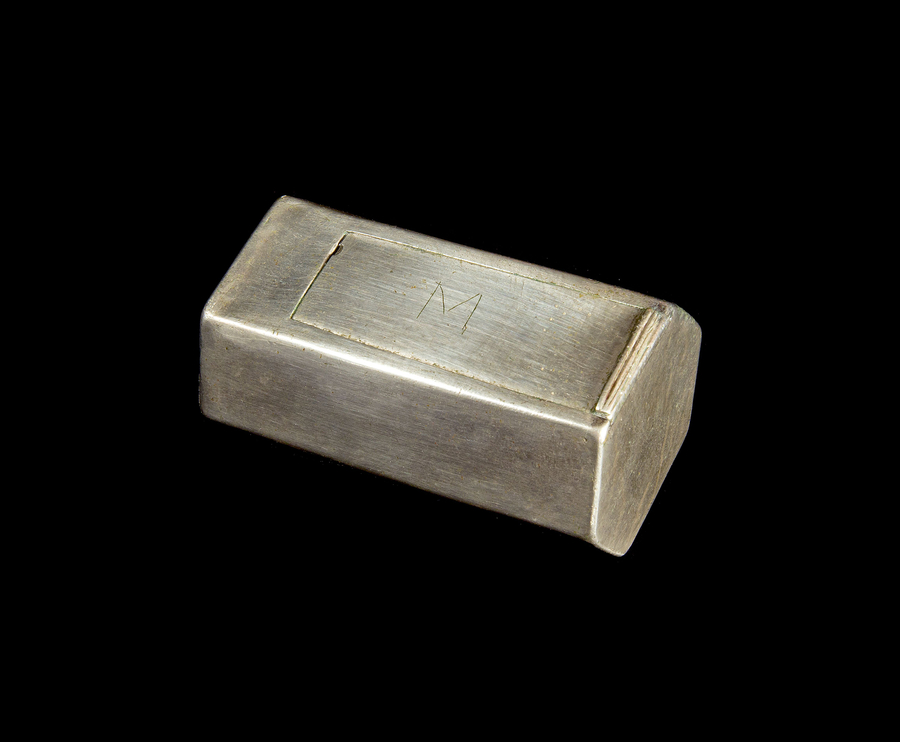 There is an M incised on the top opening of a shiny, metallic object. The object is an elongated, hexogonal cannister.