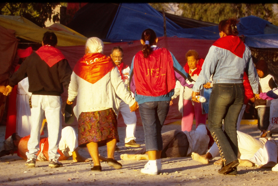 Photograph of people rolling on the floor during a pilgrimage. Others standing are wearing red capes. 