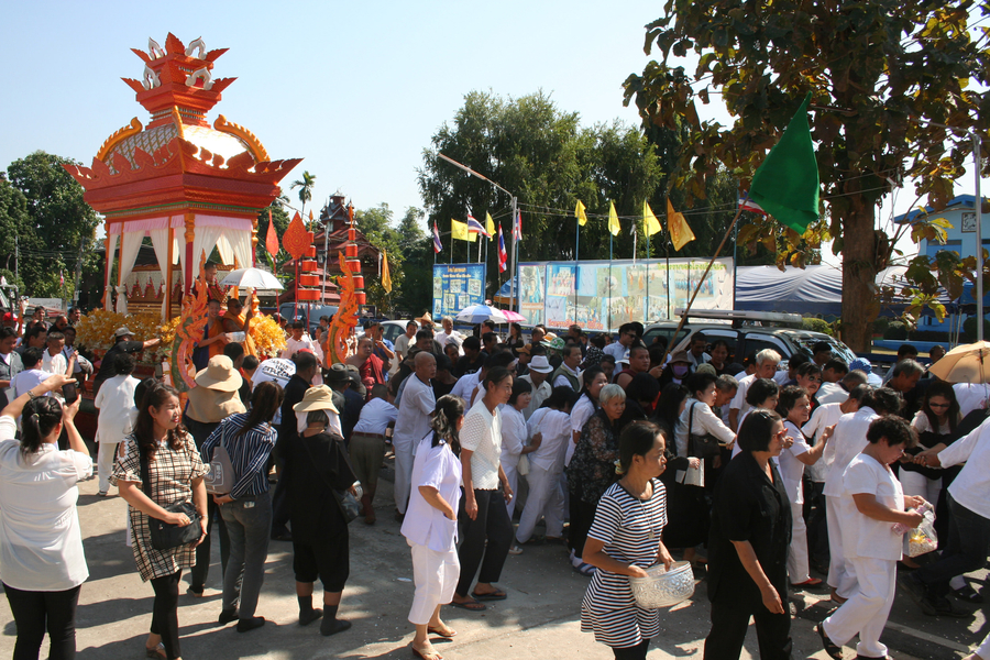 Thai people of different genders and ages accompany a massive gold prasat sop cremation structure as it processes down a street. The people are largely outfitted in white. 