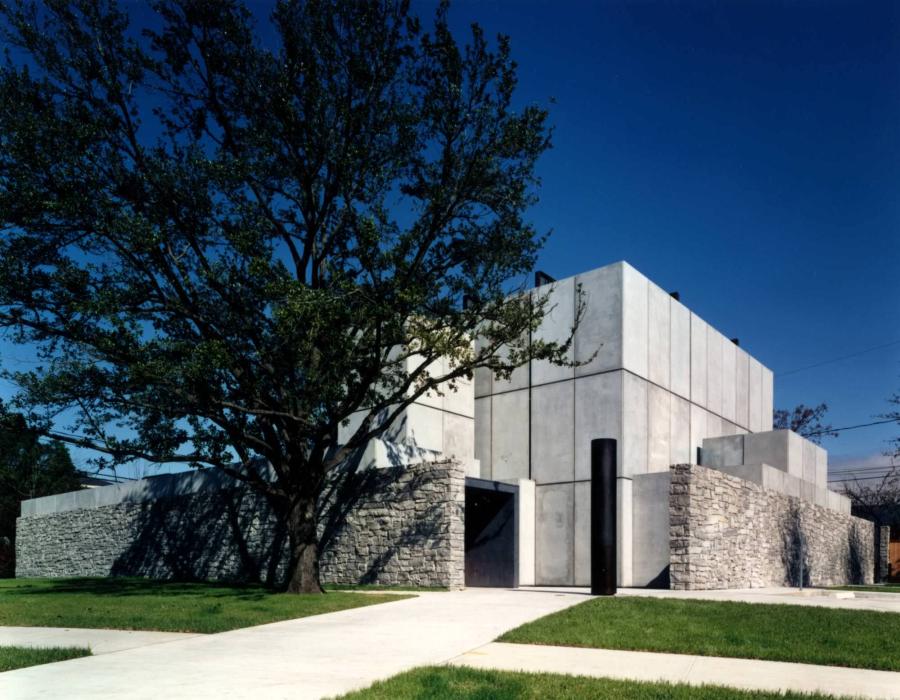 A photo captures a spare, windowless, and rectilinear limestone building against a bright blue sky and behind a lush tree. A tall stone wall encircles the building on three sides but leaves open the dark black entrance to the building.
