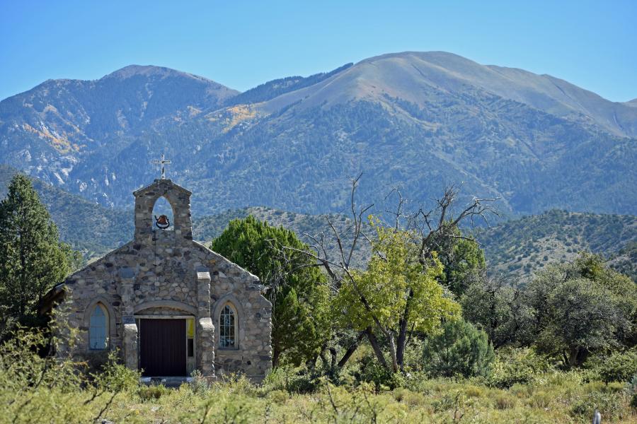 Large mountains dwarf a stone church. Its simple facade consists of two windows and stone pilasters that frame a doorway. Atop the church is a slight bell-gable and a small, faded white cross. Lush landscape abounds around the building.