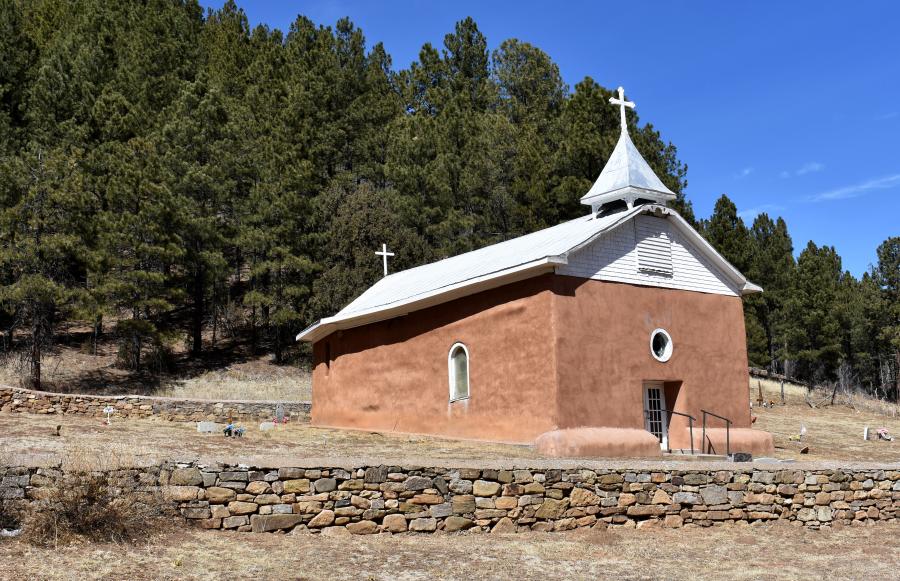 A one room sandstone chapel is set in the side of a hill and surrounded by a stone wall. Its earthen facade has a single circular window. The white gabled roof has a spire and white cross on top. Another white cross is visible on the far end of the roof.