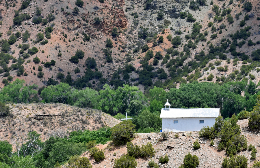 Viewed at a distance, a rectangular church is nestled in a tree-covered hillside. The white building has a gabled roof and a short bell tower, which is topped by a cross. Another larger cross stands in front of the entire building.