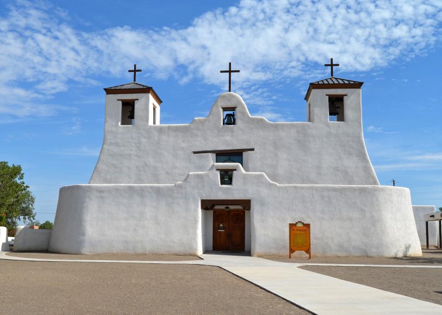 A white abode church rises up from behind a short wall that is connected to it. The church has two cross-topped spires that prominently display bells. A middle, sloped bell-gable shows off a third bell. This echoes a smaller bell-gable on the wall below.