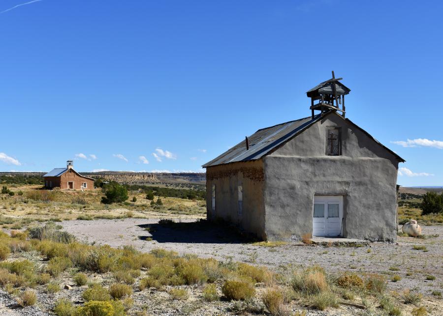 A slightly dilapidated, gray adobe church is topped with a very short and decaying wooden bell tower. The facade consists solely of a wooden cross in a slight recess of the gable. Past the church stands another structure, marked by a cross in the ground.