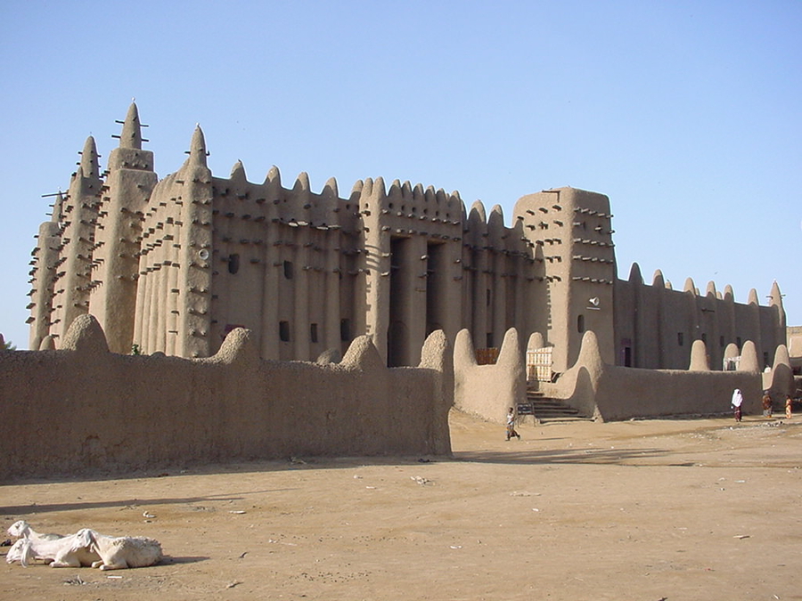 The smooth exterior of an adobe mosque is decorated with sticks of palm that jut out all over its surface. The mosque has a large trapezoidal shape with high walls that are topped by a series of cone-shaped protrusions.