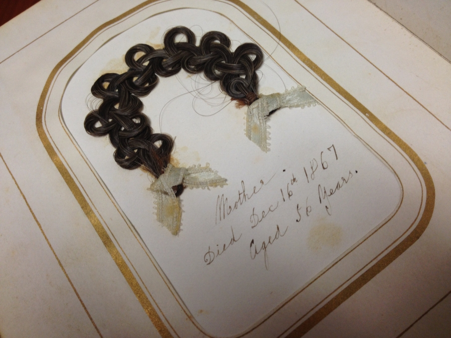 A gold border encircles a braided piece of hair arranged in an arc on a book page. Bows are tied on the hair tips. Handwritten script below reads, "Mother. Died Dec. 16th 1867. Aged 56 years."
