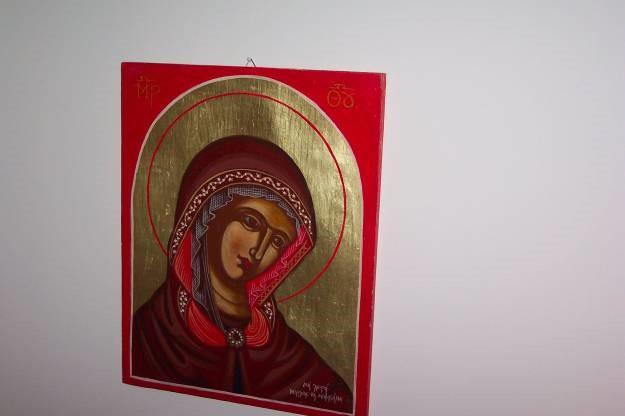 A painting of a light-skinned Mary with a large, oblong disk of gold leaf light behind her head is painted on a red background. She has large stylized eyes, red lips, and wears a burgundy cloak. 
