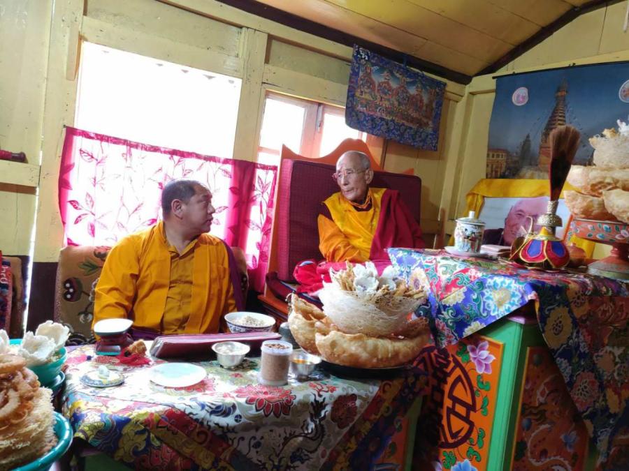 Two South Asian monks dressed in yellow and red robes sit on cushioned chairs behind colorful tables laden with food. One is older and sat on a higher chair and the other is younger and sat lower.