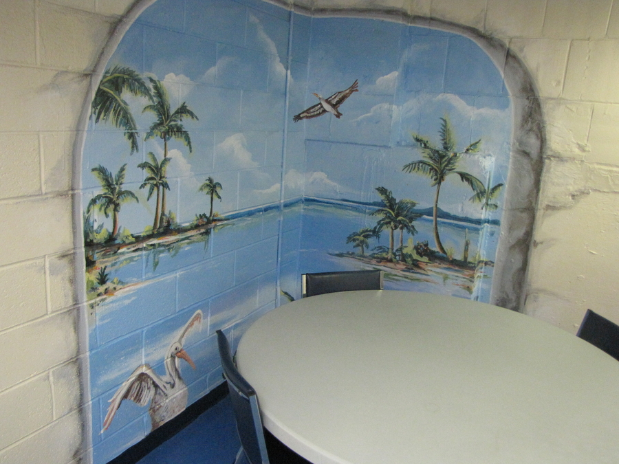 A mural of an ocean expanse with palm trees on small islands and flying seabirds is painted over the corner of a white brick wall. A white plastic table is positioned in this corner niche and covers part of the mural. 