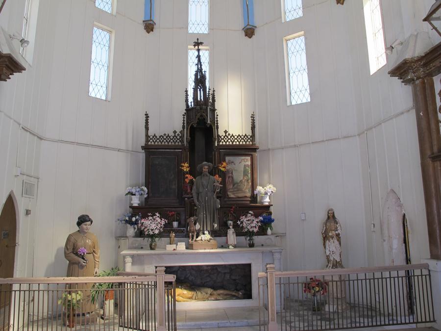 A white altar with an ornate wooden altarpiece stands in a white architectural space. A sculpture of Christ entombed lies in the altar base. A robed figure with a dog stands atop the altar among smaller flowers and statuettes.