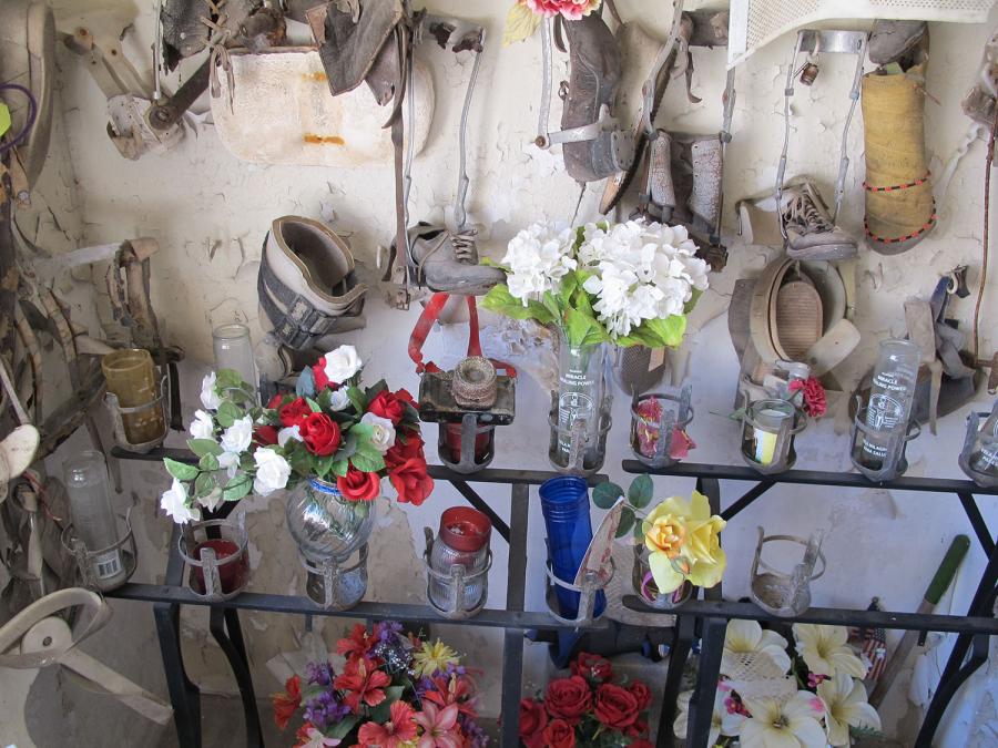 Dust-covered shoes and metal braces hang from a white wall above a metal frame. The frame holds candle and vase offerings.