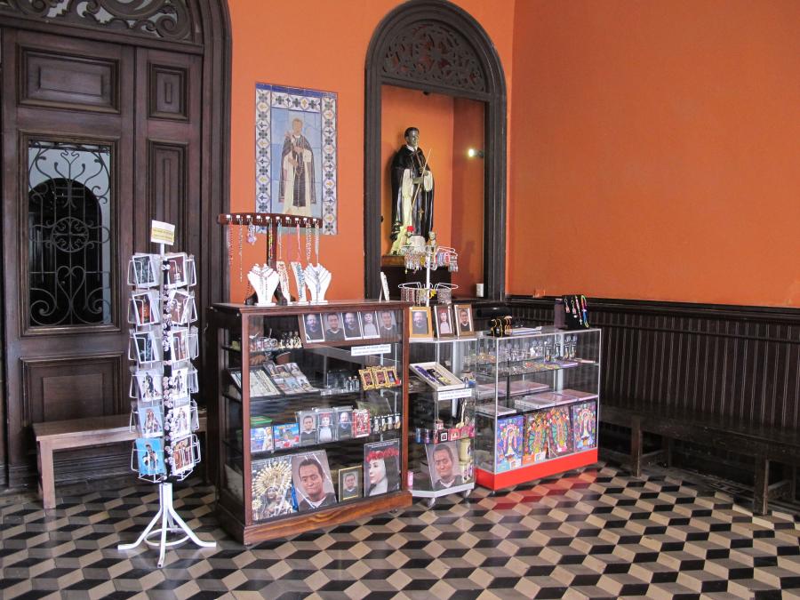 Three glass cases and a small floor spinner contain postcards and posters of saints' digital portraits as well as jewelry for sale. The souvenir shop sits in an orange-colored interior.
