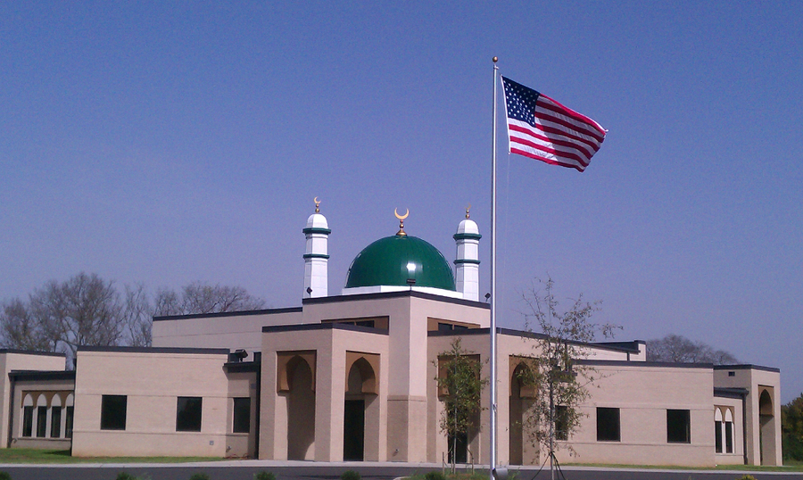 A flat-roofed, tan mosque has simple, square windows and pointed archways. A green dome with white minarets tops the building. An American flag stands in front of the complex.